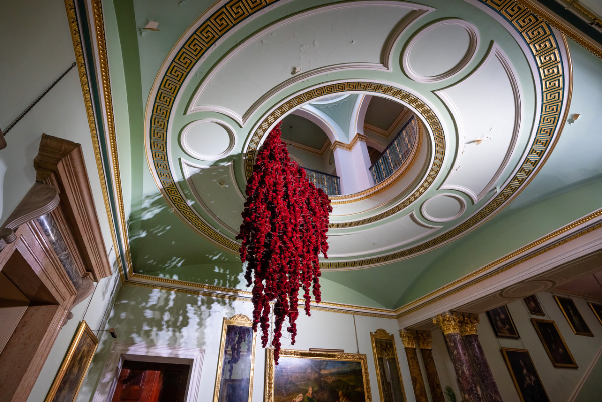 Gathering

site specific installation

Tatton Park Mansion 2019

silk velvet, reclaimed military barbed wire

Susie MacMurray has an uncanny knack of going to the heart of the matter. Her work is somehow immediately evocative, reeling in the viewer to contemplate a sense of place, a feeling of loss, and a desire to touch.  Her unusual combinations of materials and subject matter, in site-specific installations, are always intriguing and poetic. They provoke an interaction with the viewer, which draws on both physical and cultural associations.

In 'Gathering,' designed specifically to hang from the circular piercing under the rotunda of the staircase hall at Tatton Park, MacMurray returns to her constant theme of the ephemeral nature of life. To create this installation, she has worked regularly with small groups of volunteers, to hand sew tiny, luxurious, red velvet, ball-like elements, which she has then assembled to cascade in different configurations from the edge of the opening. The overall effect is of an eerie, dripping canopy of red velvet Spanish moss.

The luxury and softness of the deep red silk velvet, is impaled by protruding lengths of barbed wire, used by the MoD to train soldiers going to the Falklands, Kosovo and Afghanistan. The use of a material associated with conflict can be read as alluding to the narrative of cancer as a battle. This bringing together of such diametrically opposed materials lies at the core of MacMurray's approach. She enjoys "the conversations between not always expected combinations of materials, or materials in contexts in which you are not expecting to see them." Such juxtapositions are simultaneously appealing and disturbing. MacMurray works with and through materials, using them as a way of thinking about and expressing more conceptual ideas.

MacMurray is no stranger to loss; her husband died very young and very rapidly of cancer. In 'Gathering,' she examines this experience. The red silk she perceives as sensual, positive, bodily and precious, expressing love and desire and the living of life to the full. Yet the velvet impaled by the barbed wire is strangely beautiful and evocative. "Your life," she explains, "gets pierced by this sudden ordeal." The hospice helps by gathering people in its fold. Indeed many of the volunteers involved in the making of 'Gathering,' have been helped by the Hospice in this enfolding, supportive way, which MacMurray expresses in the gathering together of the red velvet elements.

MacMurray uses all manner of unlikely objects from rubber tubing, to barbed wire, to hair and wax, via a range of textiles and cords, combining them in strange assortments, to make them things of beauty and resonance. Her work is both solid and ethereal. She looks at the ephemeral nature of life. At life and loss, the soft, imperfect, human body that despite its imperfections is so glorious. She entices the visitor into contemplating the fragility of life, but also its joys.

Corrine Julius

June 2019

Susie MacMurray was invited to create this work to both celebrate and highlight the 20th anniversary of the Art Fair, which was established in 1999 to raise funds for the East Cheshire Hospice.

To purchase a piece of Gathering visit <a href='https://www.gatheringattatton.com/store'>gatheringattatton</a>

All funds raised go to East Cheshire Hospice.