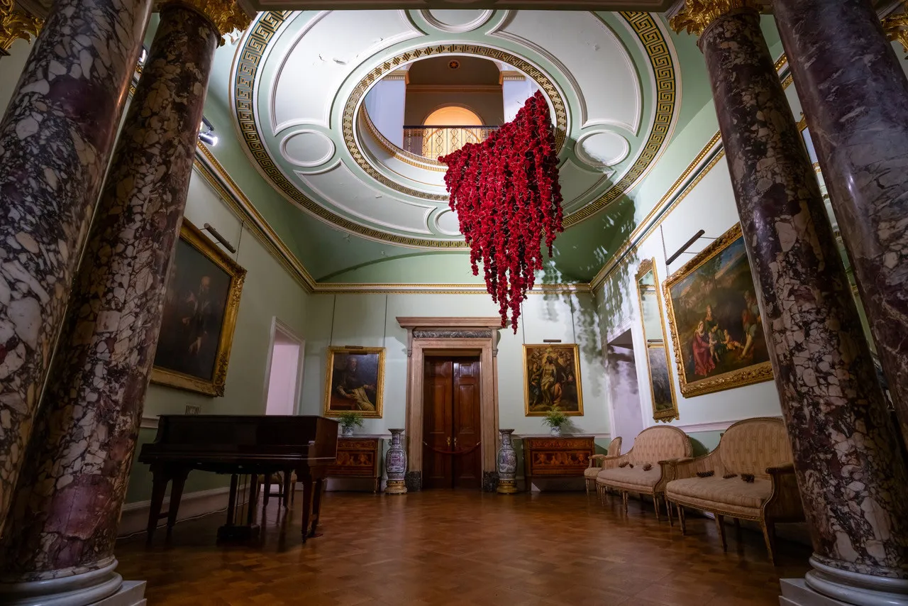 Gathering

site specific installation

Tatton Park Mansion 2019

silk velvet, reclaimed military barbed wire

Susie MacMurray has an uncanny knack of going to the heart of the matter. Her work is somehow immediately evocative, reeling in the viewer to contemplate a sense of place, a feeling of loss, and a desire to touch.  Her unusual combinations of materials and subject matter, in site-specific installations, are always intriguing and poetic. They provoke an interaction with the viewer, which draws on both physical and cultural associations.

In 'Gathering,' designed specifically to hang from the circular piercing under the rotunda of the staircase hall at Tatton Park, MacMurray returns to her constant theme of the ephemeral nature of life. To create this installation, she has worked regularly with small groups of volunteers, to hand sew tiny, luxurious, red velvet, ball-like elements, which she has then assembled to cascade in different configurations from the edge of the opening. The overall effect is of an eerie, dripping canopy of red velvet Spanish moss.

The luxury and softness of the deep red silk velvet, is impaled by protruding lengths of barbed wire, used by the MoD to train soldiers going to the Falklands, Kosovo and Afghanistan. The use of a material associated with conflict can be read as alluding to the narrative of cancer as a battle. This bringing together of such diametrically opposed materials lies at the core of MacMurray's approach. She enjoys "the conversations between not always expected combinations of materials, or materials in contexts in which you are not expecting to see them." Such juxtapositions are simultaneously appealing and disturbing. MacMurray works with and through materials, using them as a way of thinking about and expressing more conceptual ideas.

MacMurray is no stranger to loss; her husband died very young and very rapidly of cancer. In 'Gathering,' she examines this experience. The red silk she perceives as sensual, positive, bodily and precious, expressing love and desire and the living of life to the full. Yet the velvet impaled by the barbed wire is strangely beautiful and evocative. "Your life," she explains, "gets pierced by this sudden ordeal." The hospice helps by gathering people in its fold. Indeed many of the volunteers involved in the making of 'Gathering,' have been helped by the Hospice in this enfolding, supportive way, which MacMurray expresses in the gathering together of the red velvet elements.

MacMurray uses all manner of unlikely objects from rubber tubing, to barbed wire, to hair and wax, via a range of textiles and cords, combining them in strange assortments, to make them things of beauty and resonance. Her work is both solid and ethereal. She looks at the ephemeral nature of life. At life and loss, the soft, imperfect, human body that despite its imperfections is so glorious. She entices the visitor into contemplating the fragility of life, but also its joys.

Corrine Julius

June 2019

Susie MacMurray was invited to create this work to both celebrate and highlight the 20th anniversary of the Art Fair, which was established in 1999 to raise funds for the East Cheshire Hospice.

To purchase a piece of Gathering visit <a href='https://www.gatheringattatton.com/store'>gatheringattatton</a>

All funds raised go to East Cheshire Hospice.