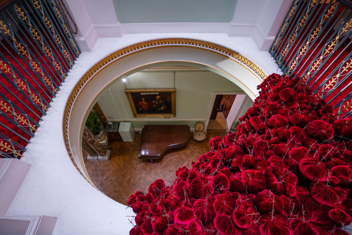 Gathering

site specific installation

Tatton Park Mansion 2019

silk velvet, reclaimed military barbed wire

Susie MacMurray has an uncanny knack of going to the heart of the matter. Her work is somehow immediately evocative, reeling in the viewer to contemplate a sense of place, a feeling of loss, and a desire to touch.  Her unusual combinations of materials and subject matter, in site-specific installations, are always intriguing and poetic. They provoke an interaction with the viewer, which draws on both physical and cultural associations.

In 'Gathering,' designed specifically to hang from the circular piercing under the rotunda of the staircase hall at Tatton Park, MacMurray returns to her constant theme of the ephemeral nature of life. To create this installation, she has worked regularly with small groups of volunteers, to hand sew tiny, luxurious, red velvet, ball-like elements, which she has then assembled to cascade in different configurations from the edge of the opening. The overall effect is of an eerie, dripping canopy of red velvet Spanish moss.

The luxury and softness of the deep red silk velvet, is impaled by protruding lengths of barbed wire, used by the MoD to train soldiers going to the Falklands, Kosovo and Afghanistan. The use of a material associated with conflict can be read as alluding to the narrative of cancer as a battle. This bringing together of such diametrically opposed materials lies at the core of MacMurray's approach. She enjoys "the conversations between not always expected combinations of materials, or materials in contexts in which you are not expecting to see them." Such juxtapositions are simultaneously appealing and disturbing. MacMurray works with and through materials, using them as a way of thinking about and expressing more conceptual ideas.

MacMurray is no stranger to loss; her husband died very young and very rapidly of cancer. In 'Gathering,' she examines this experience. The red silk she perceives as sensual, positive, bodily and precious, expressing love and desire and the living of life to the full. Yet the velvet impaled by the barbed wire is strangely beautiful and evocative. "Your life," she explains, "gets pierced by this sudden ordeal." The hospice helps by gathering people in its fold. Indeed many of the volunteers involved in the making of 'Gathering,' have been helped by the Hospice in this enfolding, supportive way, which MacMurray expresses in the gathering together of the red velvet elements.

MacMurray uses all manner of unlikely objects from rubber tubing, to barbed wire, to hair and wax, via a range of textiles and cords, combining them in strange assortments, to make them things of beauty and resonance. Her work is both solid and ethereal. She looks at the ephemeral nature of life. At life and loss, the soft, imperfect, human body that despite its imperfections is so glorious. She entices the visitor into contemplating the fragility of life, but also its joys.

Corrine Julius

June 2019

Susie MacMurray was invited to create this work to both celebrate and highlight the 20th anniversary of the Art Fair, which was established in 1999 to raise funds for the East Cheshire Hospice.

To purchase a piece of Gathering visit <a href='https://www.gatheringattatton.com/store'>gatheringattatton</a>

All funds raised go to East Cheshire Hospice.