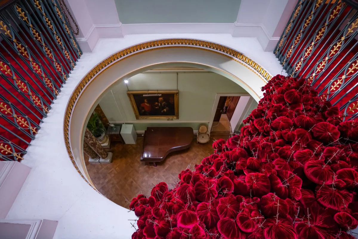 Gathering

site specific installation

Tatton Park Mansion 2019

silk velvet, reclaimed military barbed wire

Susie MacMurray has an uncanny knack of going to the heart of the matter. Her work is somehow immediately evocative, reeling in the viewer to contemplate a sense of place, a feeling of loss, and a desire to touch.  Her unusual combinations of materials and subject matter, in site-specific installations, are always intriguing and poetic. They provoke an interaction with the viewer, which draws on both physical and cultural associations.

In 'Gathering,' designed specifically to hang from the circular piercing under the rotunda of the staircase hall at Tatton Park, MacMurray returns to her constant theme of the ephemeral nature of life. To create this installation, she has worked regularly with small groups of volunteers, to hand sew tiny, luxurious, red velvet, ball-like elements, which she has then assembled to cascade in different configurations from the edge of the opening. The overall effect is of an eerie, dripping canopy of red velvet Spanish moss.

The luxury and softness of the deep red silk velvet, is impaled by protruding lengths of barbed wire, used by the MoD to train soldiers going to the Falklands, Kosovo and Afghanistan. The use of a material associated with conflict can be read as alluding to the narrative of cancer as a battle. This bringing together of such diametrically opposed materials lies at the core of MacMurray's approach. She enjoys "the conversations between not always expected combinations of materials, or materials in contexts in which you are not expecting to see them." Such juxtapositions are simultaneously appealing and disturbing. MacMurray works with and through materials, using them as a way of thinking about and expressing more conceptual ideas.

MacMurray is no stranger to loss; her husband died very young and very rapidly of cancer. In 'Gathering,' she examines this experience. The red silk she perceives as sensual, positive, bodily and precious, expressing love and desire and the living of life to the full. Yet the velvet impaled by the barbed wire is strangely beautiful and evocative. "Your life," she explains, "gets pierced by this sudden ordeal." The hospice helps by gathering people in its fold. Indeed many of the volunteers involved in the making of 'Gathering,' have been helped by the Hospice in this enfolding, supportive way, which MacMurray expresses in the gathering together of the red velvet elements.

MacMurray uses all manner of unlikely objects from rubber tubing, to barbed wire, to hair and wax, via a range of textiles and cords, combining them in strange assortments, to make them things of beauty and resonance. Her work is both solid and ethereal. She looks at the ephemeral nature of life. At life and loss, the soft, imperfect, human body that despite its imperfections is so glorious. She entices the visitor into contemplating the fragility of life, but also its joys.

Corrine Julius

June 2019

Susie MacMurray was invited to create this work to both celebrate and highlight the 20th anniversary of the Art Fair, which was established in 1999 to raise funds for the East Cheshire Hospice.

To purchase a piece of Gathering visit <a href='https://www.gatheringattatton.com/store'>gatheringattatton</a>

All funds raised go to East Cheshire Hospice.