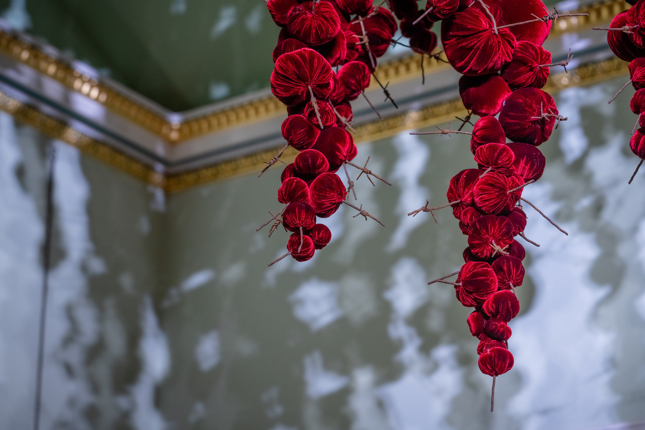 Gathering

site specific installation

Tatton Park Mansion 2019

silk velvet, reclaimed military barbed wire

Susie MacMurray has an uncanny knack of going to the heart of the matter. Her work is somehow immediately evocative, reeling in the viewer to contemplate a sense of place, a feeling of loss, and a desire to touch.  Her unusual combinations of materials and subject matter, in site-specific installations, are always intriguing and poetic. They provoke an interaction with the viewer, which draws on both physical and cultural associations.

In 'Gathering,' designed specifically to hang from the circular piercing under the rotunda of the staircase hall at Tatton Park, MacMurray returns to her constant theme of the ephemeral nature of life. To create this installation, she has worked regularly with small groups of volunteers, to hand sew tiny, luxurious, red velvet, ball-like elements, which she has then assembled to cascade in different configurations from the edge of the opening. The overall effect is of an eerie, dripping canopy of red velvet Spanish moss.

The luxury and softness of the deep red silk velvet, is impaled by protruding lengths of barbed wire, used by the MoD to train soldiers going to the Falklands, Kosovo and Afghanistan. The use of a material associated with conflict can be read as alluding to the narrative of cancer as a battle. This bringing together of such diametrically opposed materials lies at the core of MacMurray's approach. She enjoys "the conversations between not always expected combinations of materials, or materials in contexts in which you are not expecting to see them." Such juxtapositions are simultaneously appealing and disturbing. MacMurray works with and through materials, using them as a way of thinking about and expressing more conceptual ideas.

MacMurray is no stranger to loss; her husband died very young and very rapidly of cancer. In 'Gathering,' she examines this experience. The red silk she perceives as sensual, positive, bodily and precious, expressing love and desire and the living of life to the full. Yet the velvet impaled by the barbed wire is strangely beautiful and evocative. "Your life," she explains, "gets pierced by this sudden ordeal." The hospice helps by gathering people in its fold. Indeed many of the volunteers involved in the making of 'Gathering,' have been helped by the Hospice in this enfolding, supportive way, which MacMurray expresses in the gathering together of the red velvet elements.

MacMurray uses all manner of unlikely objects from rubber tubing, to barbed wire, to hair and wax, via a range of textiles and cords, combining them in strange assortments, to make them things of beauty and resonance. Her work is both solid and ethereal. She looks at the ephemeral nature of life. At life and loss, the soft, imperfect, human body that despite its imperfections is so glorious. She entices the visitor into contemplating the fragility of life, but also its joys.

Corrine Julius

June 2019

Susie MacMurray was invited to create this work to both celebrate and highlight the 20th anniversary of the Art Fair, which was established in 1999 to raise funds for the East Cheshire Hospice.

To purchase a piece of Gathering visit <a href='https://www.gatheringattatton.com/store'>gatheringattatton</a>

All funds raised go to East Cheshire Hospice.
